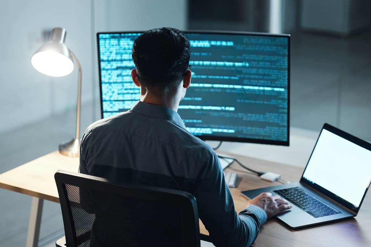 A developer seen from behind, working at a desk with a large monitor displaying code and a laptop on the side. testimonial