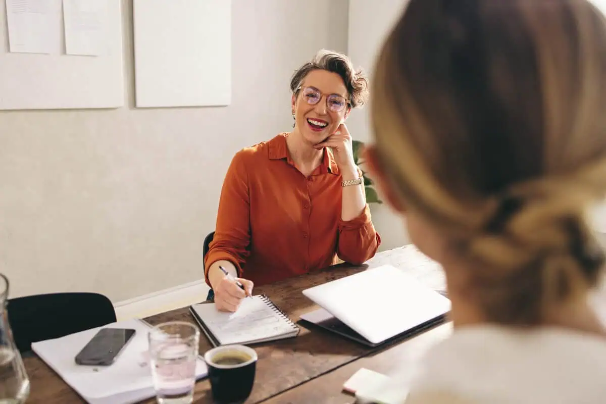 A smiling female HR expert in an orange shirt sits at a desk across from a candidate, taking notes during an interview.