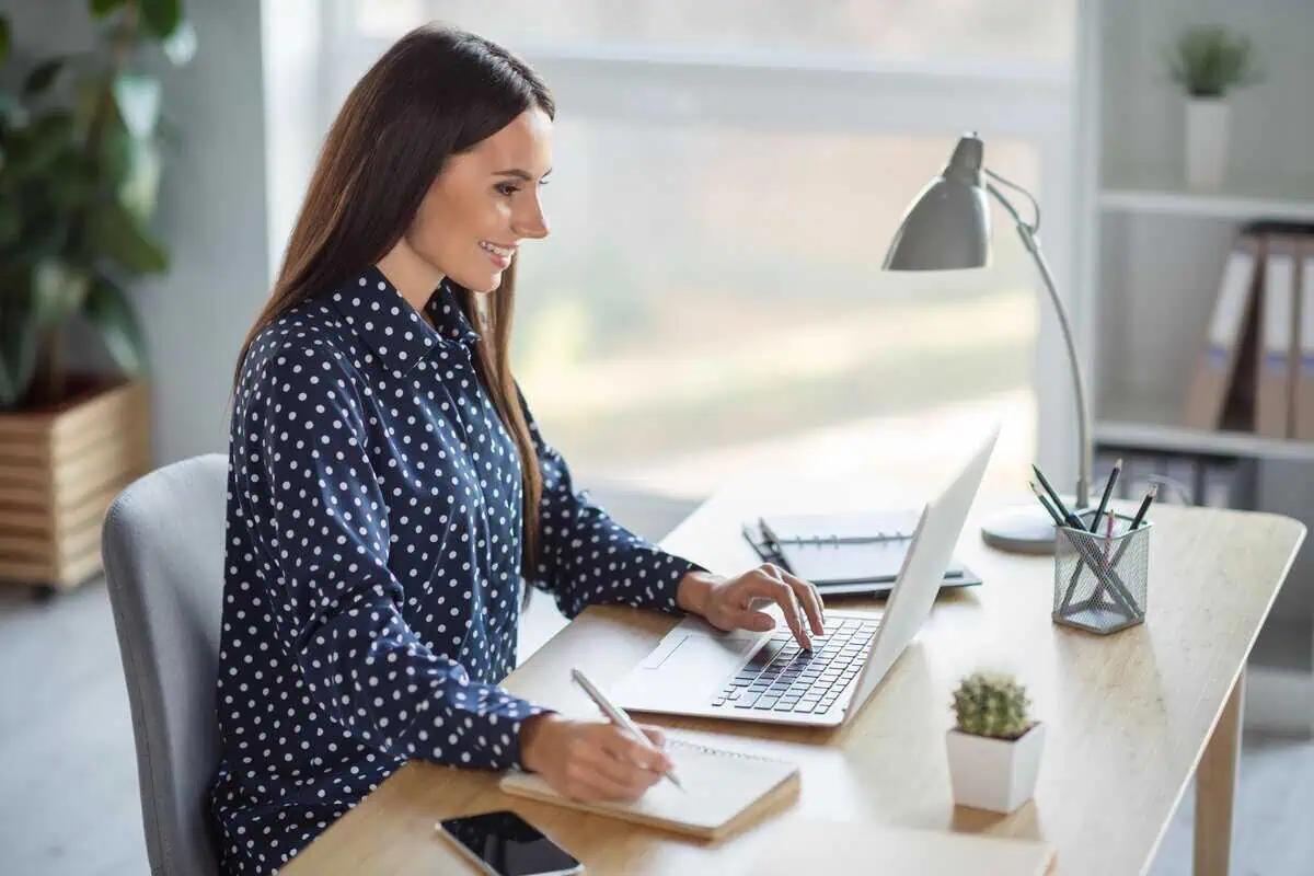 A smiling woman in a blue dotted blouse works on her laptop at a modern office desk.