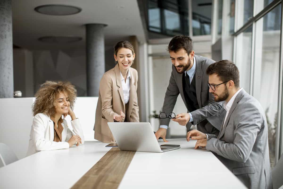 A diverse team of four professionals collaborating around a laptop in a modern office meeting room. testimonial