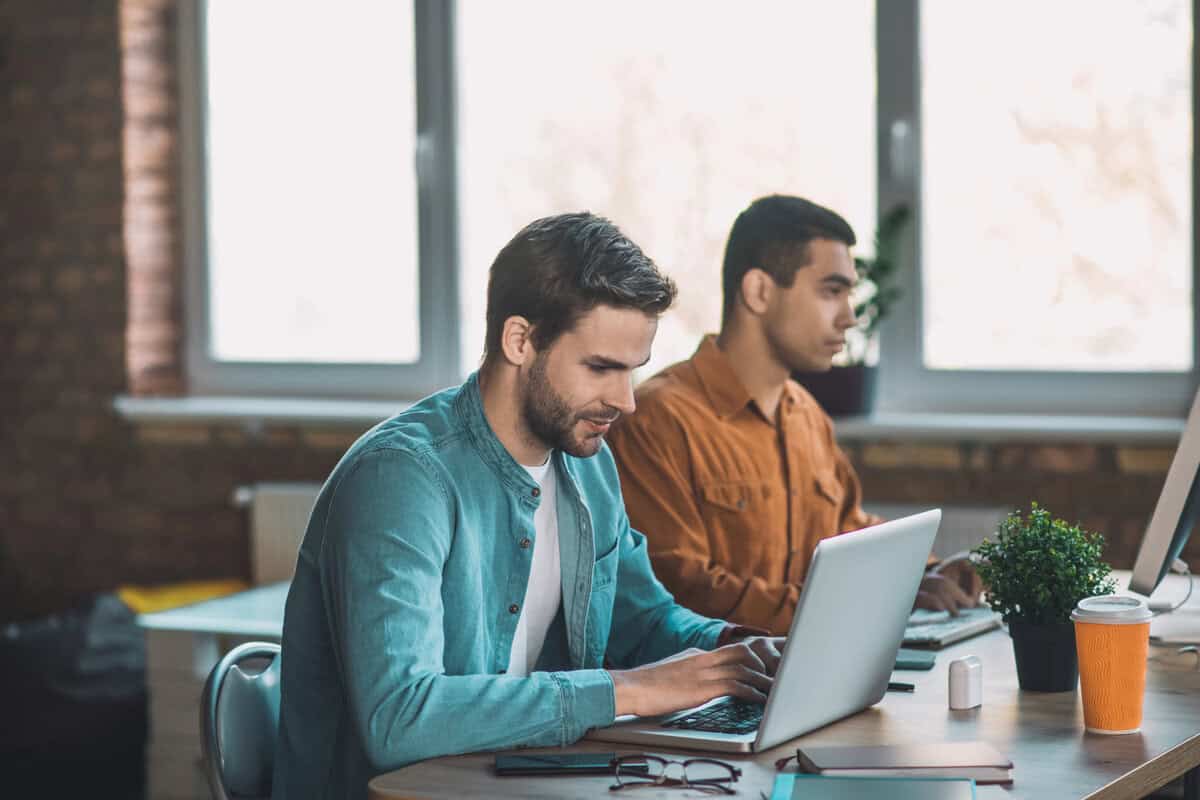 Two male developers sitting side-by-side, working on laptops in a modern office. testimonial