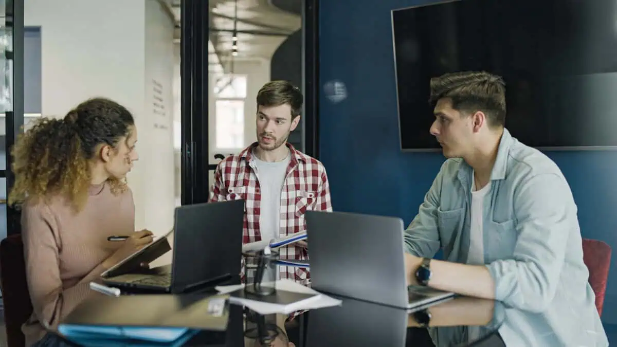 Three developers having a discussion around laptops at a meeting table.