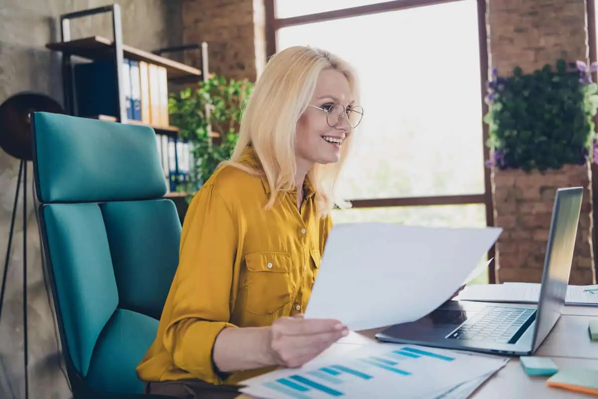 A smiling blonde woman in a yellow blouse working on a laptop and reviewing documents at a desk.