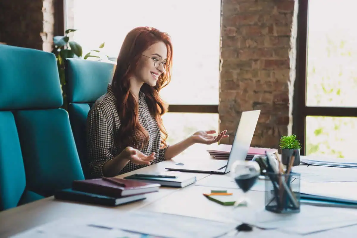 A woman with red hair and glasses smiling while working on a laptop in a modern office setting.