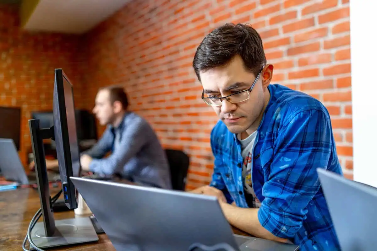 Two male developers sitting side-by-side, working on laptops in a modern office.