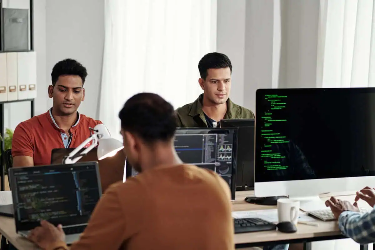 Two male developers sitting side-by-side, working on laptops in a modern office.