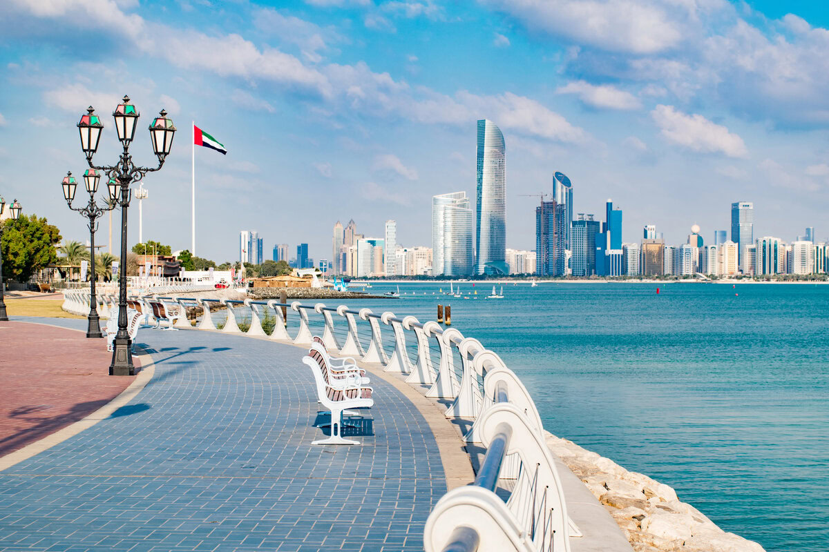 A scenic view of the Abu Dhabi skyline with a coastal walkway, street lamps, and a UAE flag. testimonial