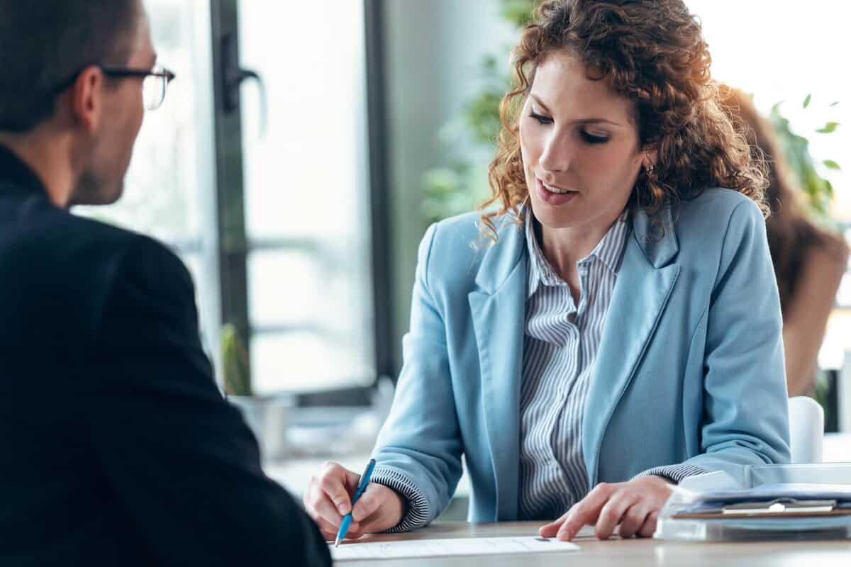 A female professional in a blue blazer points to a laptop screen while collaborating with male colleagues at a modern conference table. testimonial