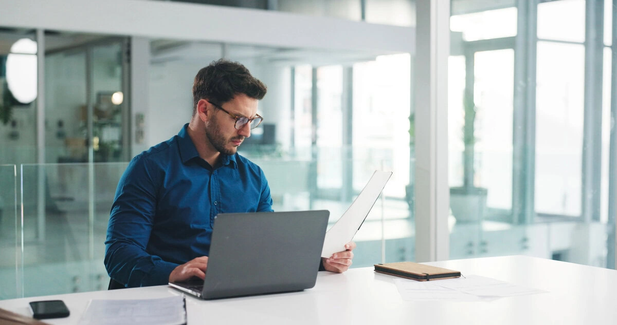 A professional man in a blue shirt working on a laptop in a bright, modern office setting.
