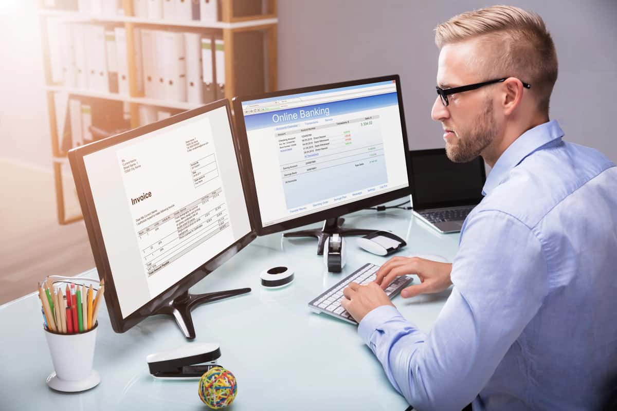 A professional working on two computer monitors, one showing an invoice and the other online banking software. testimonial
