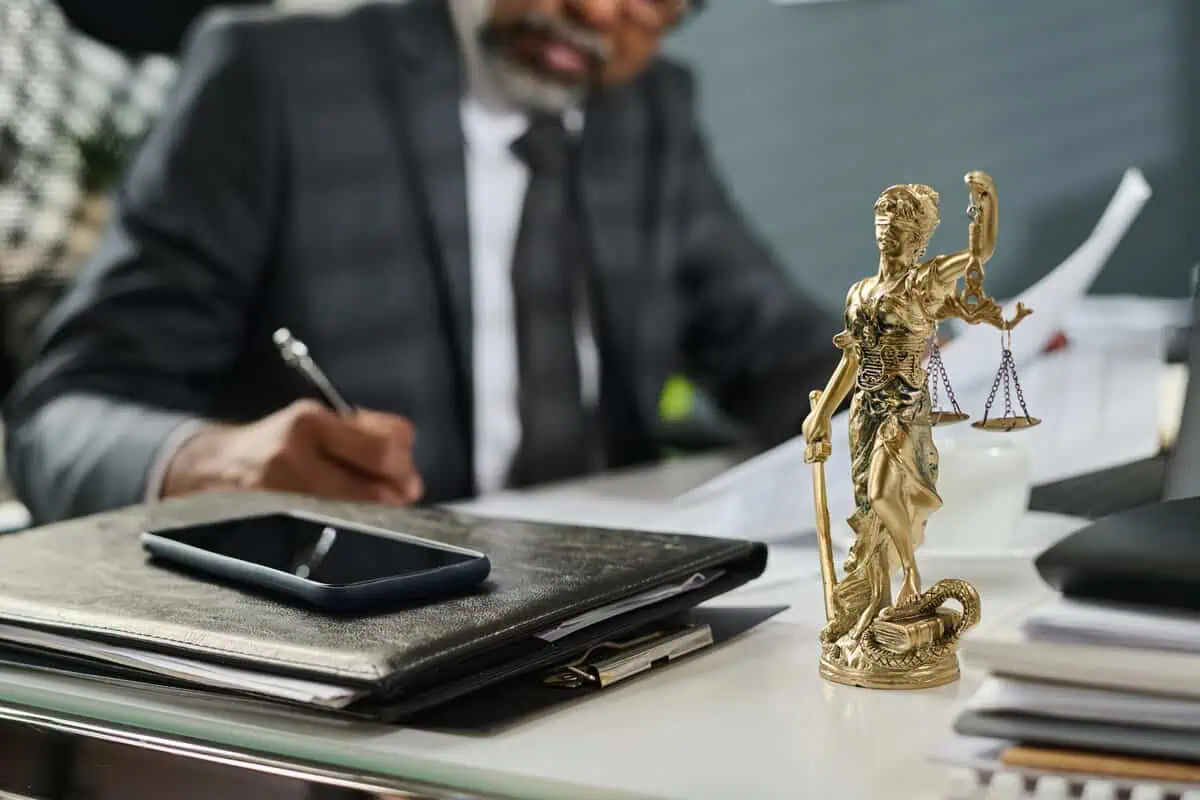 A professional at a desk signing documents with a gold Scales of Justice statue in the foreground.