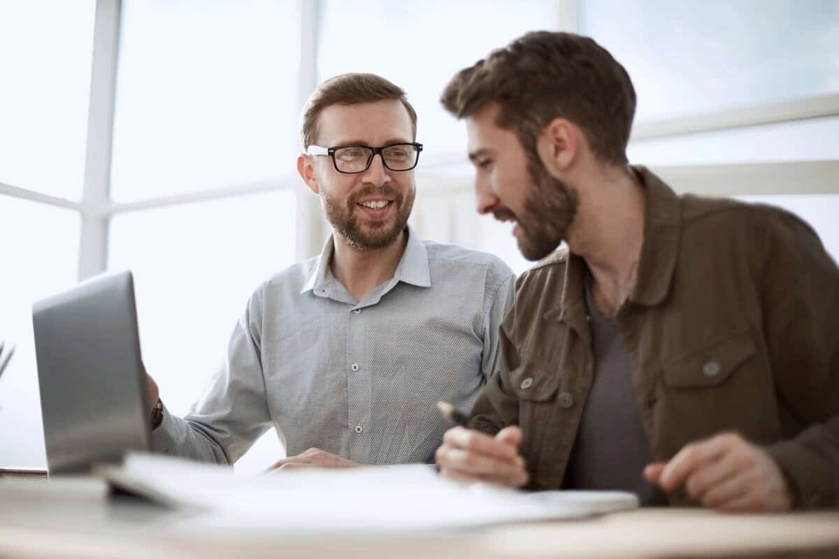 Two professional men in an office discussing a project over a laptop and documents. testimonial