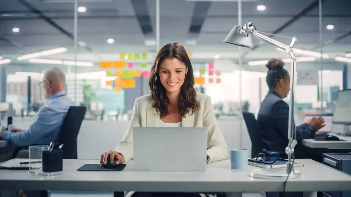 A professional woman working on a laptop in a modern office with colorful sticky notes on the glass wall behind her.